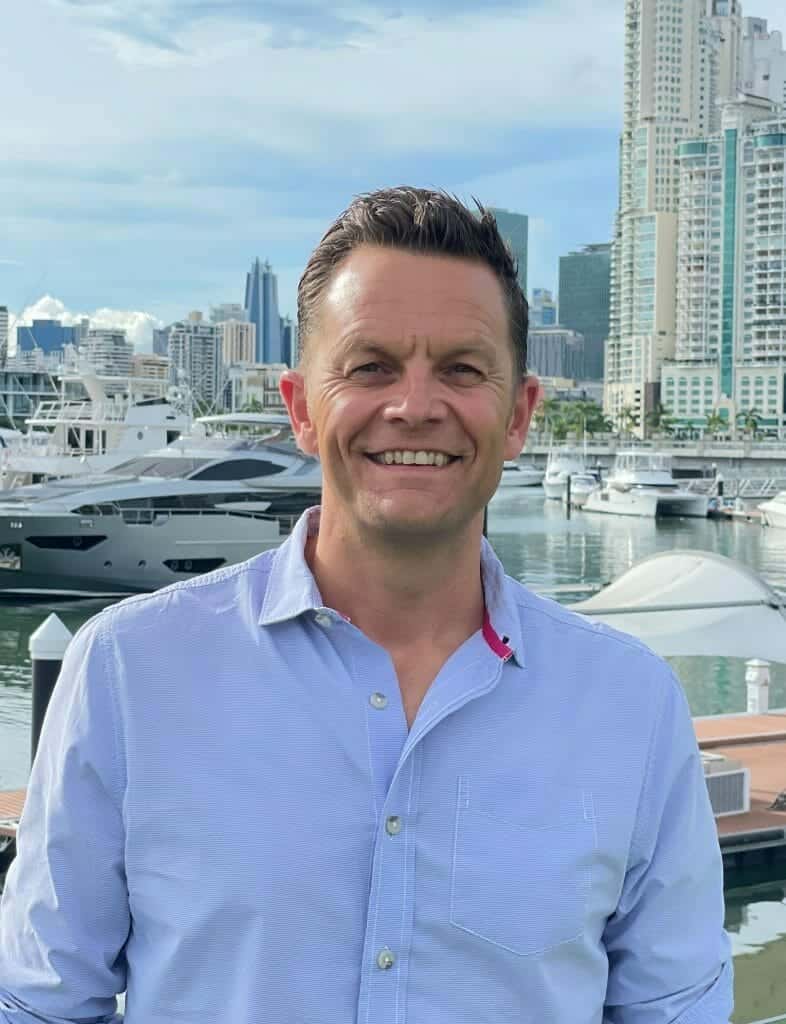 Mann in hellblauem Hemd, Klaus Happ (Geschäftsführender Gesellschafter & Gründer), lächelt in die Kamera, im Hintergrund Yachthafen und moderne Hochhäuser von Panama City.