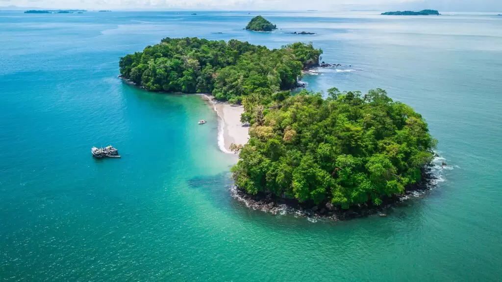 Drohnenaufnahme einer kleinen tropischen Insel bei Boca Chica in Panama mit dichtem Regenwald, weißem Sandstrand und türkisblauem Meer, daneben ankert ein Boot.